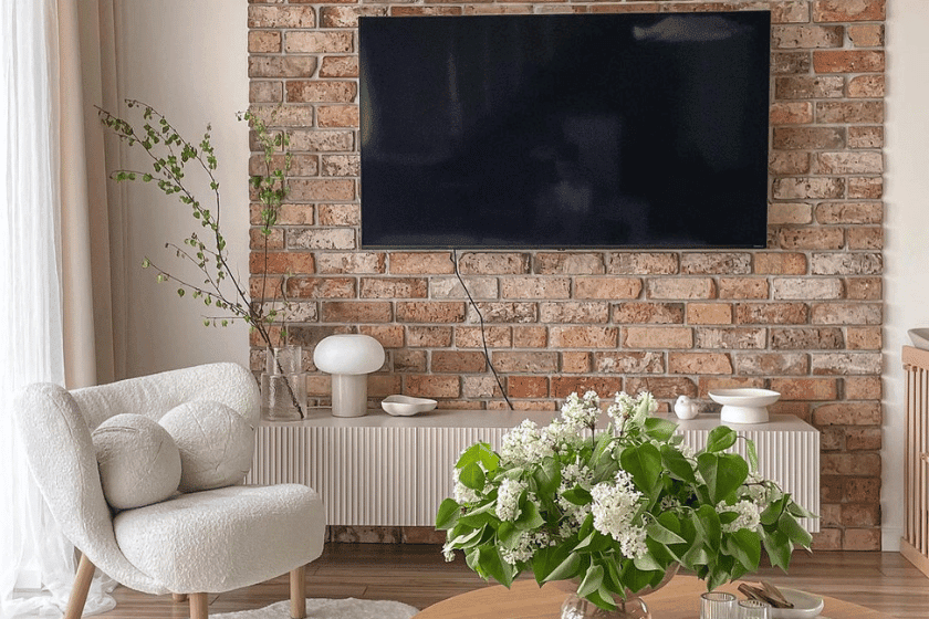 Stylowa szafka RTV z ryflowanymi frontami w jasnym salonie z ceglaną ścianą, dużym telewizorem i przytulnym fotelem boucle.
