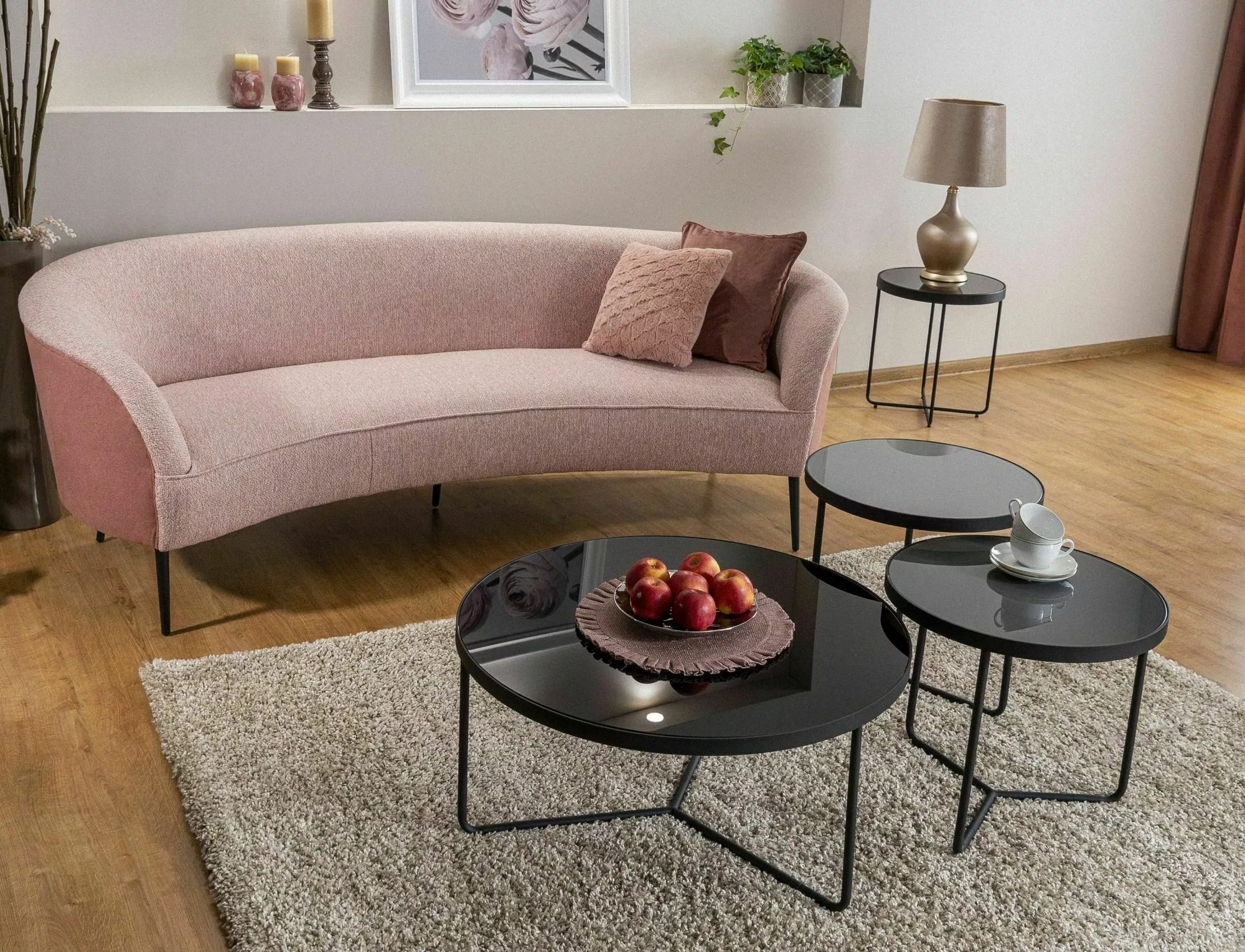 Elegancki salon z różową, welurową sofą na metalowych nóżkach i zestawem trzech okrągłych stolików kawowych z czarnym, szklanym blatem.