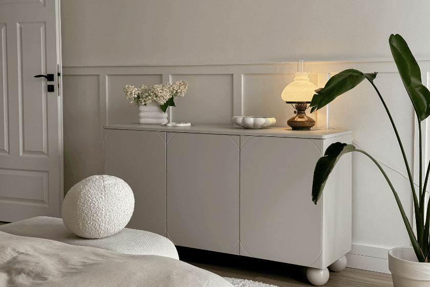 Elegancka jasna komoda na kulistych nogach w przytulnej sypialni, ozdobiona lampką, roślinami i dekoracjami w stylu minimalistycznym.