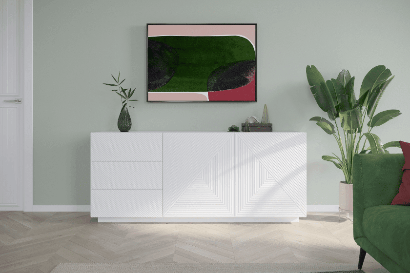 Biała komoda z geometrycznym wzorem, zielona roślina w tle, zielona sofa i kolorowa abstrakcyjna grafika na ścianie.