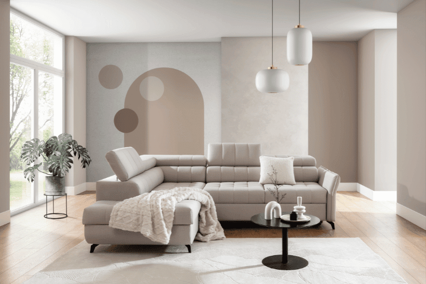 Stylowy salon z narożnikiem w odcieniu beżowym, okrągłym stołem kawowym i prostym, nowoczesnym wystrojem. W tle widoczna abstrakcyjna ściana w odcieniach beżu i ciepłych brązów.